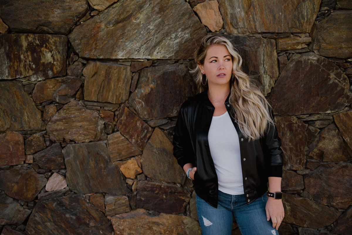 the laughing survivor alexandra stevenson stands against a stonework wall wearing a leather jacket white t and jeans looking off in the distance