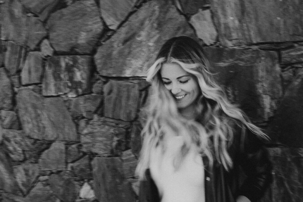 a slightly blurry laughing survivor alexandra stevensen leans against a rock wall in downtown kamloops and looks down smiling while the wind blows her long blonde hair around